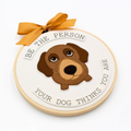 A circular embroidered wall hanging or ornament featuring a brown dachshund face in the center, surrounded by the text 'BE THE PERSON YOUR DOG THINKS YOU ARE' on a white fabric background. A mustard yellow ribbon is tied at the top, and the hoop is photographed on a clean white background.