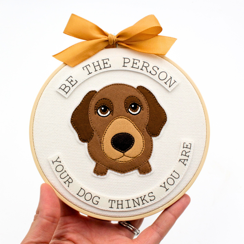 Handmade fabric hoop art piece with dog gazing upwards, surrounded by wording that says "Be the person your dog thinks you are".