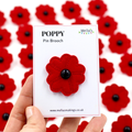 A hand holds a white card displaying a red fabric poppy pin brooch with a black center. The card is labeled 'POPPY Pin Brooch' and shows the brand 'Mella's'.