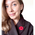 A close-up portrait of a woman wearing a bright red poppy flower brooch with a black center on her dark coat lapel.
